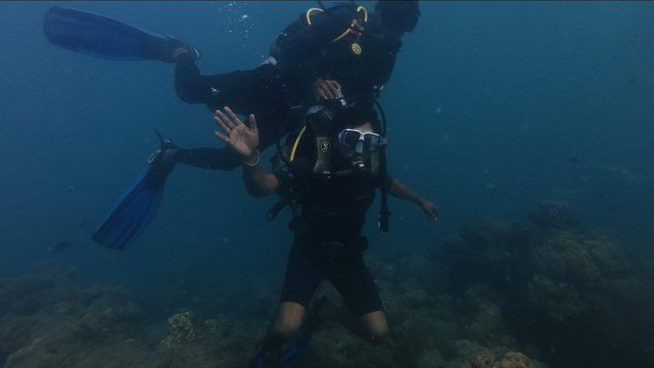 Diver waving upside-down in open blue water as a buddy with bright blue fins descends past