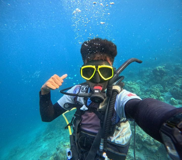 Diver's GoPro selfie giving a thumbs-up signal, reef and fish blurred behind